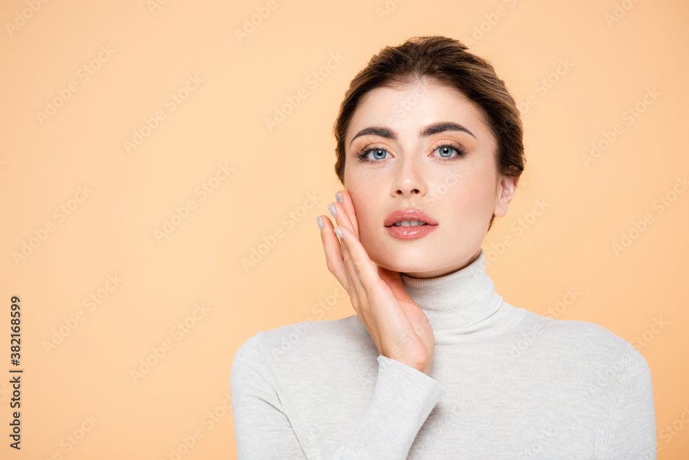 woman in turtleneck looking at camera while touching face isolated on peach woman in turtleneck looking at camera while touching face isolated on peach