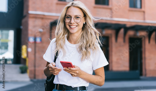 Half length portrait of cheerful caucasian female in trendy wear spending time on street using smartphone, beautiful millennial hipster girl blogger looking at camera in town holding mobile phone photo