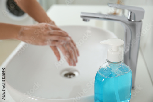 Bottle of antibacterial soap and blurred woman washing hands on background. Personal hygiene during COVID-19 pandemic