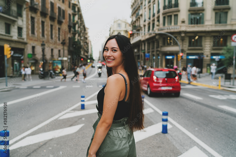Attractive caucasian tourist female with dark hair looking back at camera while walking the street. Smiling student girl wearing trendy outfit and sunglasses enjoying the first visit to European city.