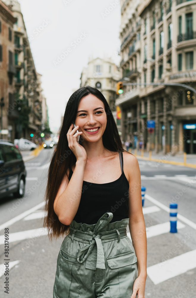 Cheerful caucasian hipster girl standing at the city street and talking on her smartphone. Young attractive woman in trendy outfit having phone conversation while standing on a city road background