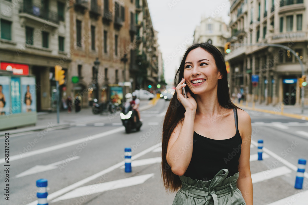 Young caucasian female is talking by the smartphone while standing on a street. A charming student girl is smiling while calling to a friend by the mobile phone. Hipster girl in trendy clothes outdoor