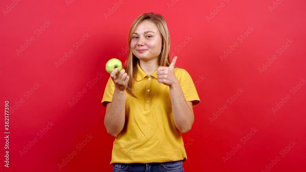 Fat and cheerful blonde woman who holds a white apple and recommends it for a healthy lifestyle, The girl in the yellow T-shirt enjoys tasty fruits rich in vitamins. Studio on a red background. Fat and cheerful blonde woman who holds a white apple and recommends it for a healthy lifestyle, The girl in the yellow T-shirt enjoys tasty fruits rich in vitamins. Studio on a red background.