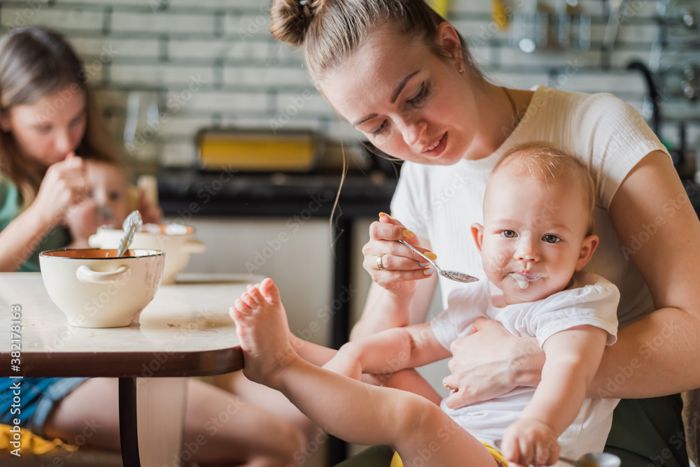 Two tired but happy mothers feed their babies milk porridge in the kitchen Two tired but happy mothers feed their babies milk porridge in the kitchen