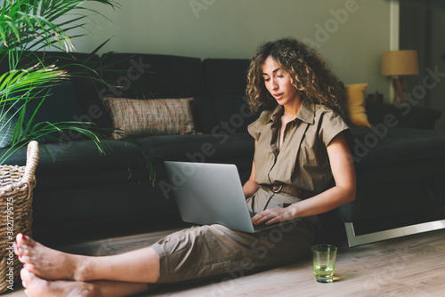 Fotografia Beautiful female in casual outfit chilling on a floor beside comfortable couch in a living room with laptop on her knees