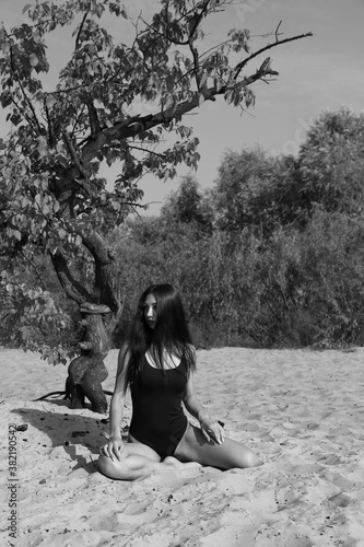 Slim long-haired young woman in bikini sitting on the sand near old tree in black and white
