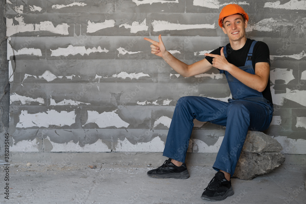 male builder dressed in work clothes and an orange mask. Sit on a stone with index fingers on an empty space