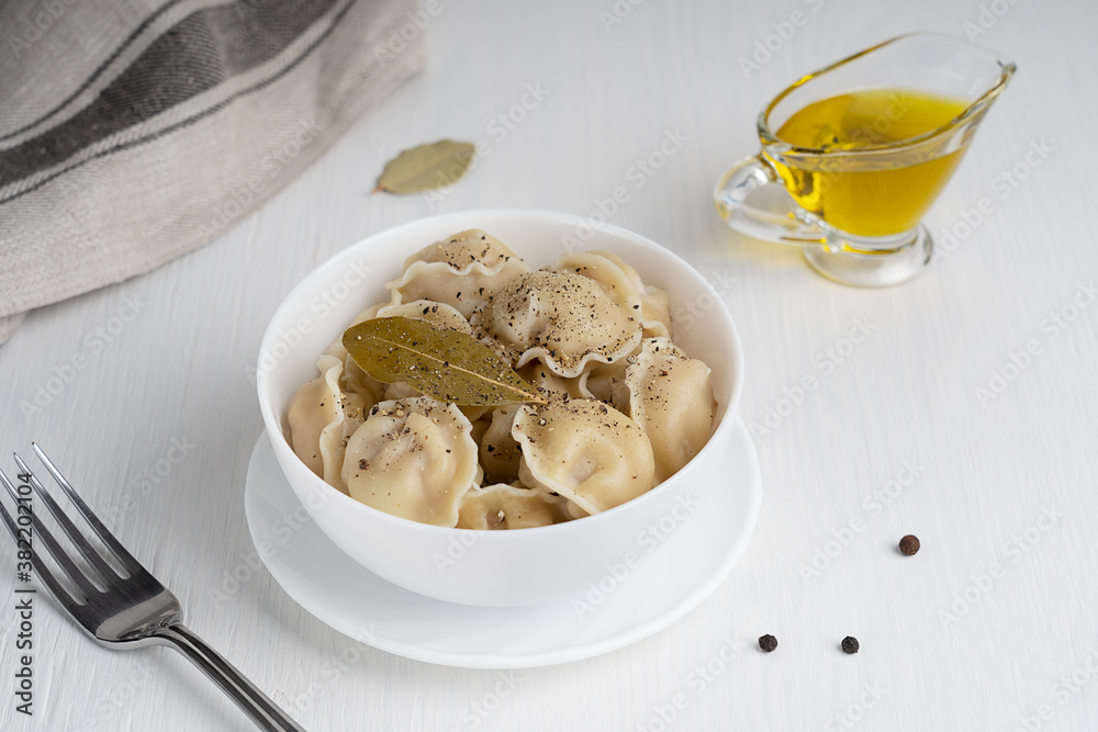 Pelmeni or dumplings of Russian cuisine made of minced meat filling wrapped in dough served in white bowl with fork with black pepper seasoning and bay leaf on white wooden background. Horizontal