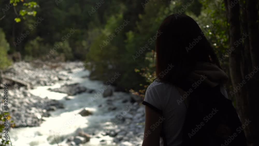 A young woman with a backpack stands and looks at the mountain river. A young woman with a backpack stands and looks at the mountain river.