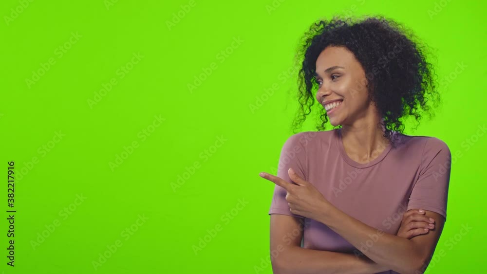 Smiling young African American woman with kinky hair wearing purple t-shirt points with forefinger to side posing on green background closeup. Check this out. Advertisement and ethnicity concept
