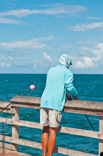 back view, medium distance of a young, adult male leaning against a wood railing, fishing off a wood pier, jutting into tropical waters of gulf of Mexico