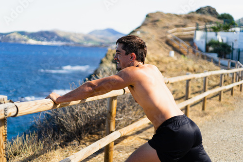 Young sportsman stretching his legs outdoors while looking at the horizon with the sea to the side.Model without shirt.