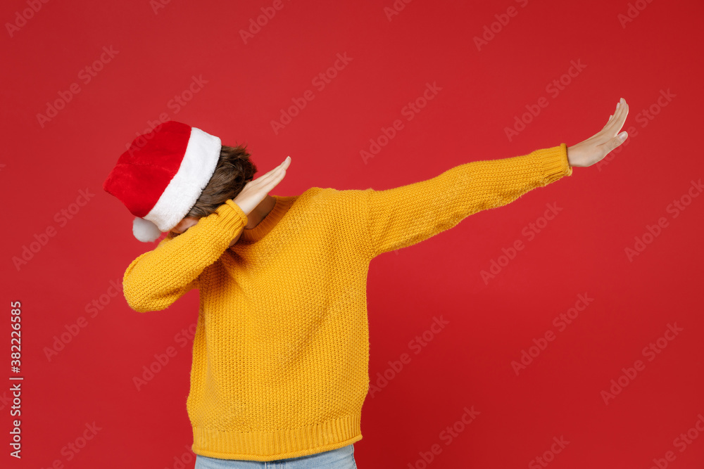 Young Santa woman in Christmas hat doing dab hip hop dance hands gesture, youth sign hiding covering face isolated on red background studio portrait. Happy New Year celebration merry holiday concept.