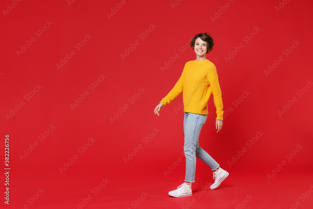 Full length side view of smiling cheerful beautiful attractive young brunette woman 20s in basic yellow sweater walking going looking camera isolated on bright red colour background studio portrait.