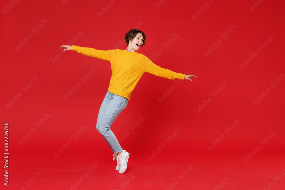 Full length side view of funny attractive young brunette woman 20s wearing basic yellow sweater dancing standing on toes spreading hands isolated on bright red colour background studio portrait.