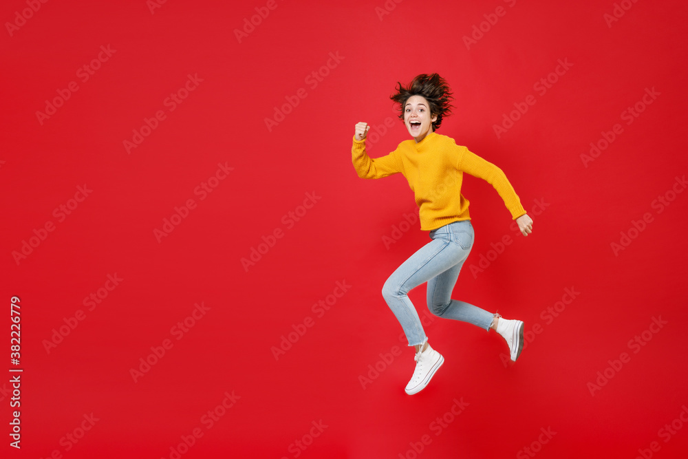 Full length side view excited surprised laughing beautiful young brunette woman 20s wearing basic casual yellow sweater jumping like running isolated on bright red colour background studio portrait.