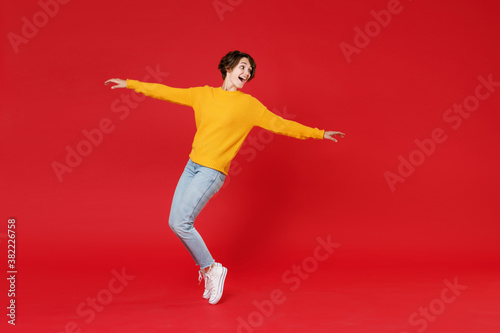 Full length side view of funny attractive young brunette woman 20s wearing basic yellow sweater dancing standing on toes spreading hands isolated on bright red colour background studio portrait.
