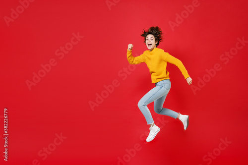 Full length side view excited surprised laughing beautiful young brunette woman 20s wearing basic casual yellow sweater jumping like running isolated on bright red colour background studio portrait.