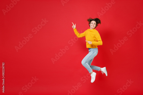 Full length side view of smiling young brunette woman 20s in basic yellow sweater jumping pointing index fingers aside on mock up copy space isolated on bright red colour background studio portrait.
