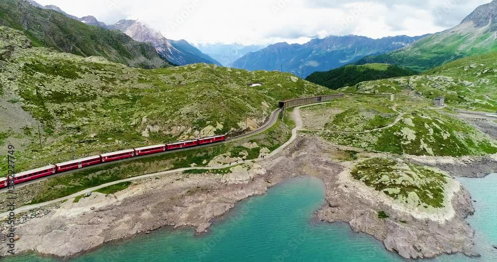 Aerial view with a drone of a red train passing near a blue lakeat the bottom of a mountain in Switzerland . Beautiful landscape under the blue sky 4K