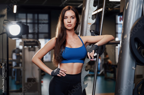 Seductive brunette sportswoman in tight sportswear with serious face posing leaning on barbell in gym.