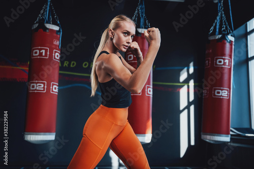 Obraz na plátně Serious slim sportswoman with blond hairs posing in fighting stance with punching bags in background in box club