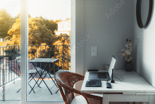 An interior of a cozy room with a desktop and a laptop on it with a USB hub and wireless graphic tablet near on the table; a glass door leads to a small balcony with a beautiful sunny view and a table photo