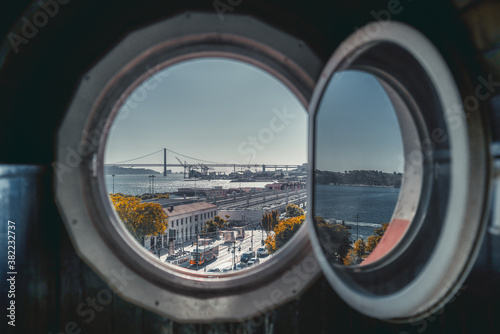 View through the round window of an antique house of Lisbon cityscape in the background with the bridge over the Tagus river, road with old trams, and modern cars on a sunny day; defocused foreground