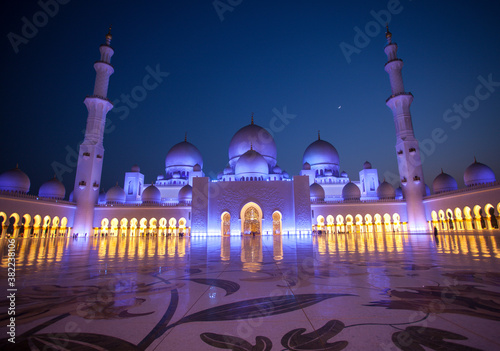 Amazing Sheikh Zayed Grand Mosque at night time, illuminated and crescent visible between two minarets. People visit the mosque for praying. Muslim mosque concept.