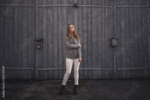 beautiful young blonde model girl. White pants. gray knitted sweater. black boots. wooden pendant on the neck in the form of a horse. On the Sunset. on a background of gray wooden gates © Andrii