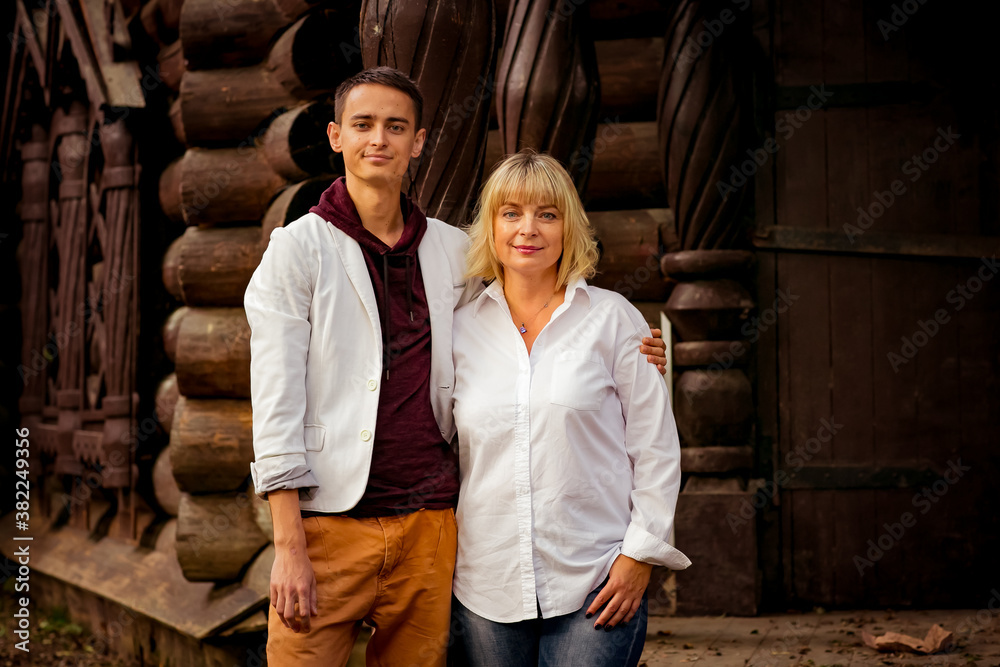 A beautiful woman,blonde,middle-aged,in a white shirt and jeans,with a large son,stands near an eco-friendly wooden house
