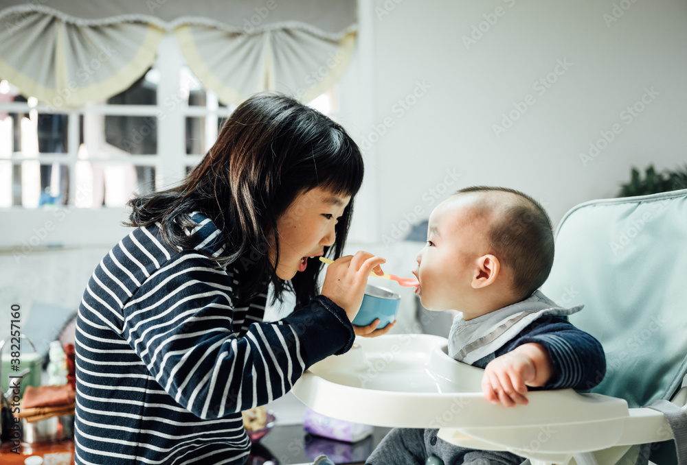Sweet girl feeding her baby boy brother with mashed vegetables in living room Sweet girl feeding her baby boy brother with mashed vegetables in living room