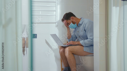 Unhappy worried businessman working on laptop wearing mask sit on the toilet at home restroom bearded wc isolation quarantine pandemic coronavirus covid-19 portrait close up slow motion