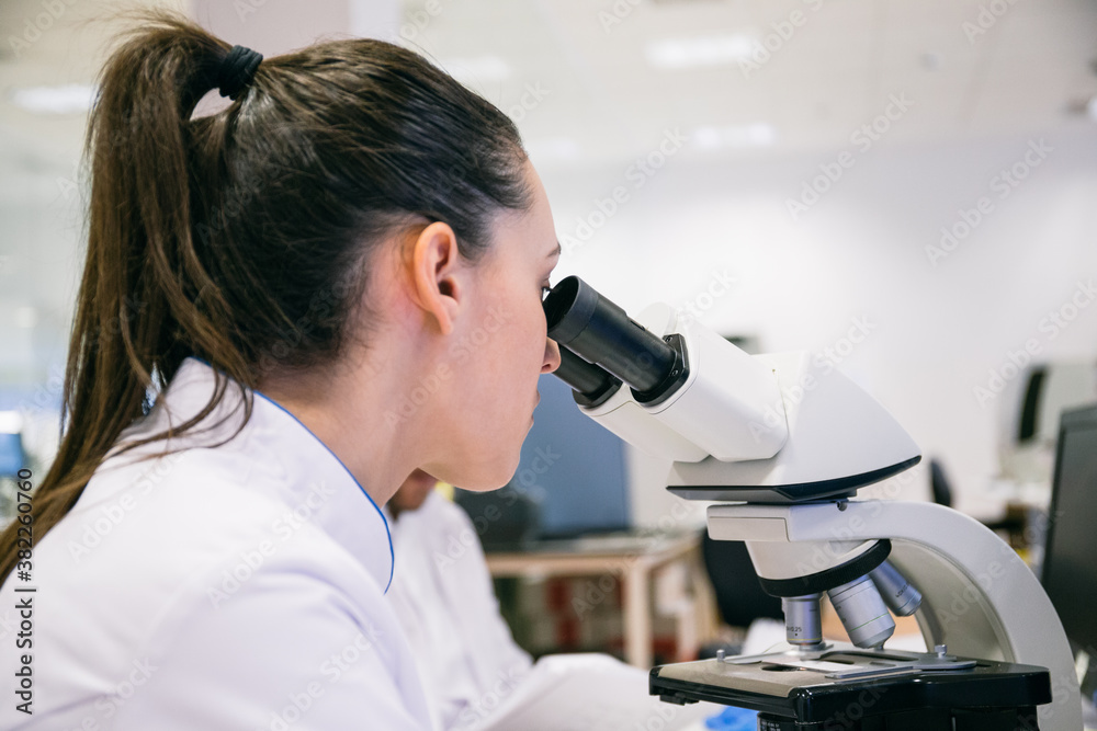 Young woman in lab robe examining samples with microscope while working in modern laboratory Young woman in lab robe examining samples with microscope while working in modern laboratory