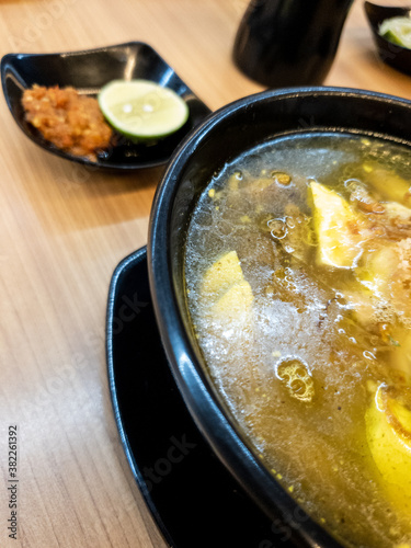 soup served in a black bowl complete with vegetables, seasonings, spoons & forks, asian style food served on a wooden table.