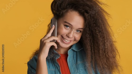 Portrait of pretty brunette girl talking with best friend on smartphone happily looking in camera isolated on yellow background