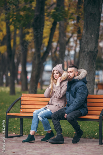 A young man and woman are resting on a bench in an autumn park. A loving couple in jackets sits on a bench in the main park