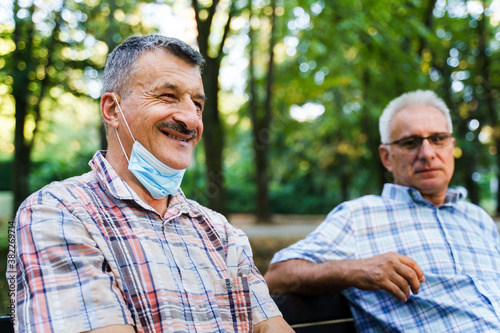 Two senior caucasian men sitting on bench in park in autumn or summer day - selective focus on man with mustaches smiling - real people leisure activity social distance concept side view copy space