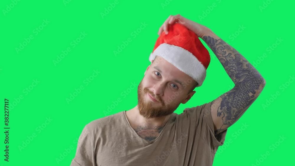 Young handsome bearded tattooed guy in green t-shirt is put on a red christmas hat on chroma key background. Isolated portrait of happy smiling man for keying Young handsome bearded tattooed guy in green t-shirt is put on a red christmas hat on chroma key background. Isolated portrait of happy smiling man for keying