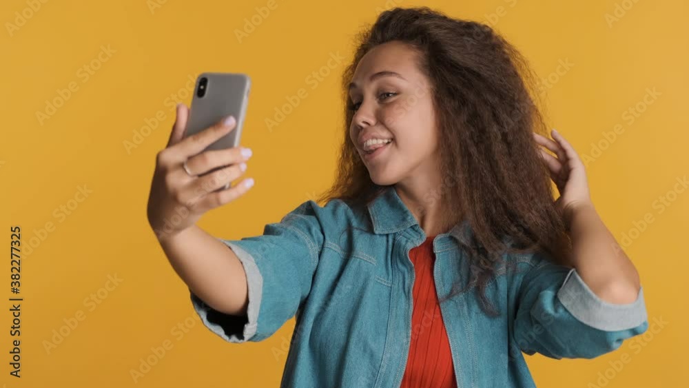 Beautiful brunette girl making selfie on smartphone and choosing photo for social network isolated on yellow background Beautiful brunette girl making selfie on smartphone and choosing photo for social network isolated on yellow background