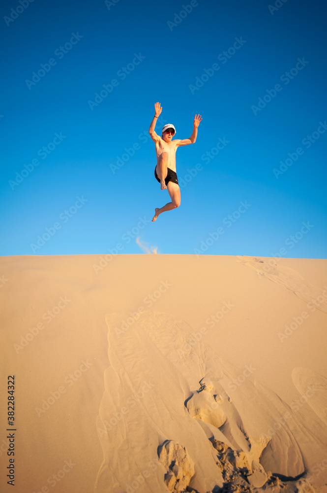 Fototapeta premium Youth jumping off a sand dune shirtless with hat and sunglasses on a clear day cloudless sky
