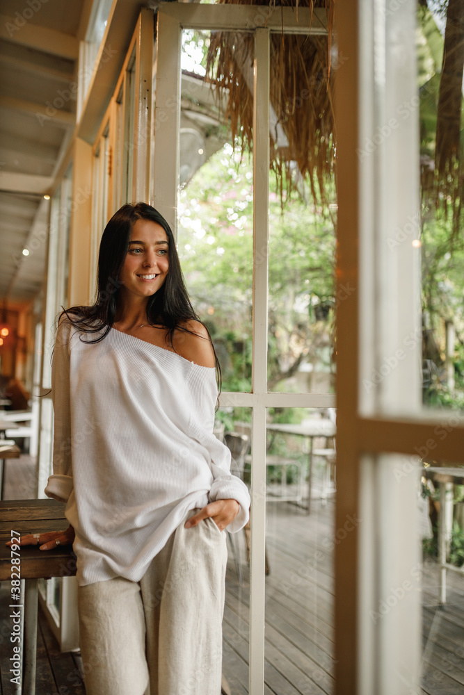 Beautiful young woman stands in front of the window in cafe and smiling