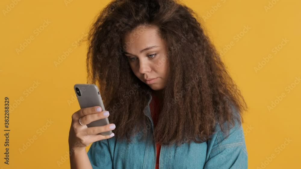 Serious brunette girl intently surfes social media on smartphone isolated on yellow background Serious brunette girl intently surfes social media on smartphone isolated on yellow background