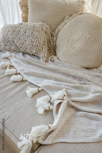 Angled closeup view of white macrame pillow and a knitted blanket lie on a beige sofa with soft focus