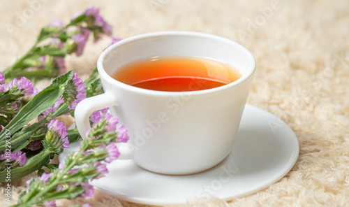 White cup with tea placed on a cream carpet. Decorated with purple flowers in the corner. 
