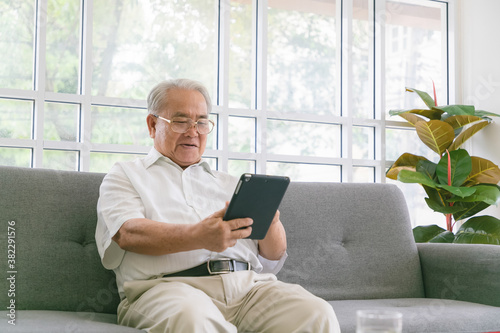 Senior asian man sitting on sofa and reading new on his tablet computer.