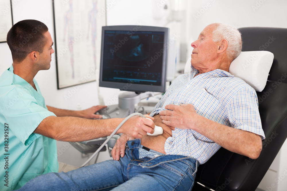 Obraz premium Young male doctor using ultrasound scan doing abdominal diagnostic to senior male patient in modern clinic
