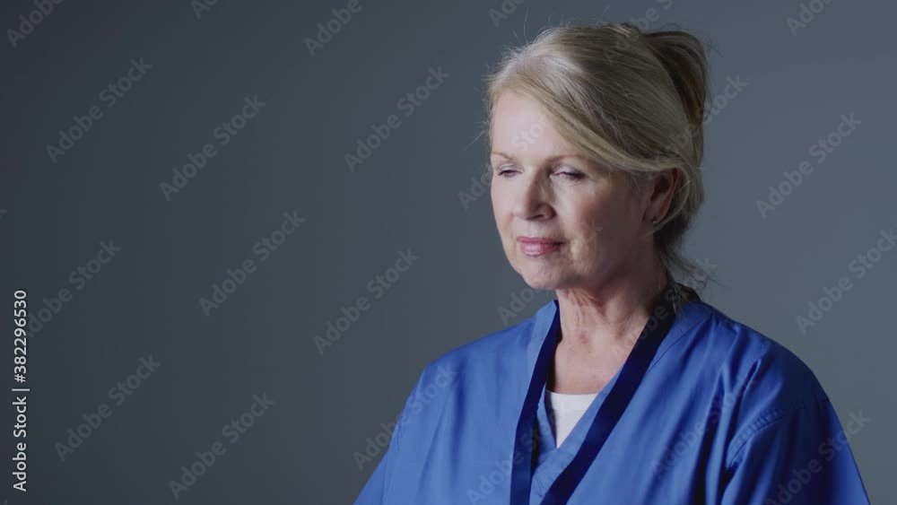 Studio portrait of smiling mature female nurse wearing scrubs against grey background - shot in slow motion Studio portrait of smiling mature female nurse wearing scrubs against grey background - shot in slow motion