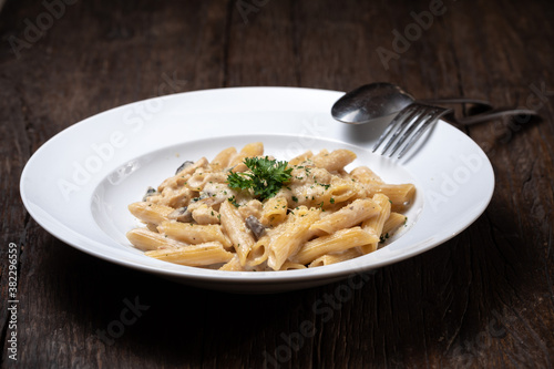 Homemade delicious pasta macaroni with white mushrooms chicken meet mushrooms in creamy cheese sauce in the white plate on the dark brown wood table. Parsley  spicy. Yellow  green.