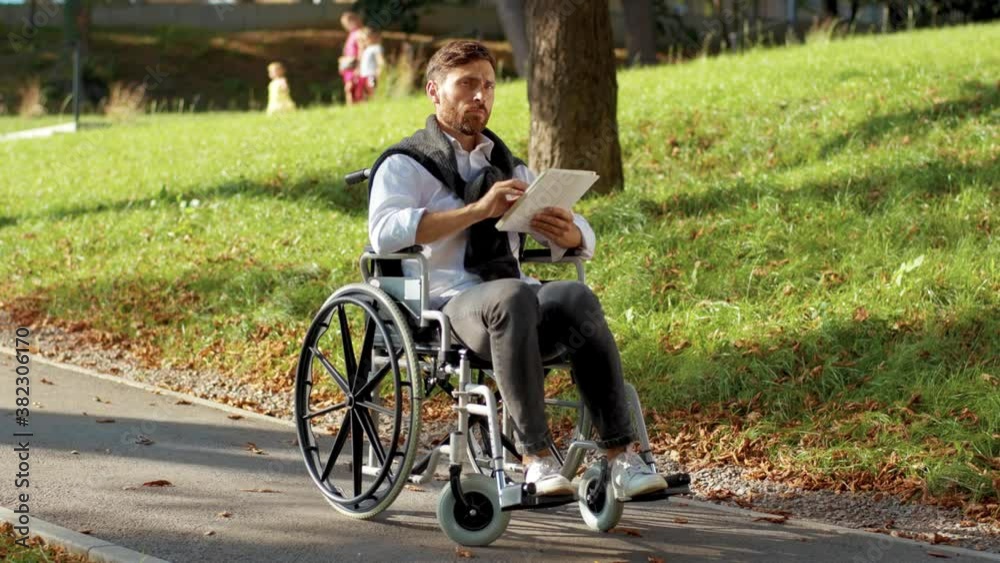Attractive young man on a wheelchair with tablet computer in park at sunlight disability medicine. Possibility handicapped handicap tablet internet. Slow motion Attractive young man on a wheelchair with tablet computer in park at sunlight disability medicine. Possibility handicapped handicap tablet internet. Slow motion